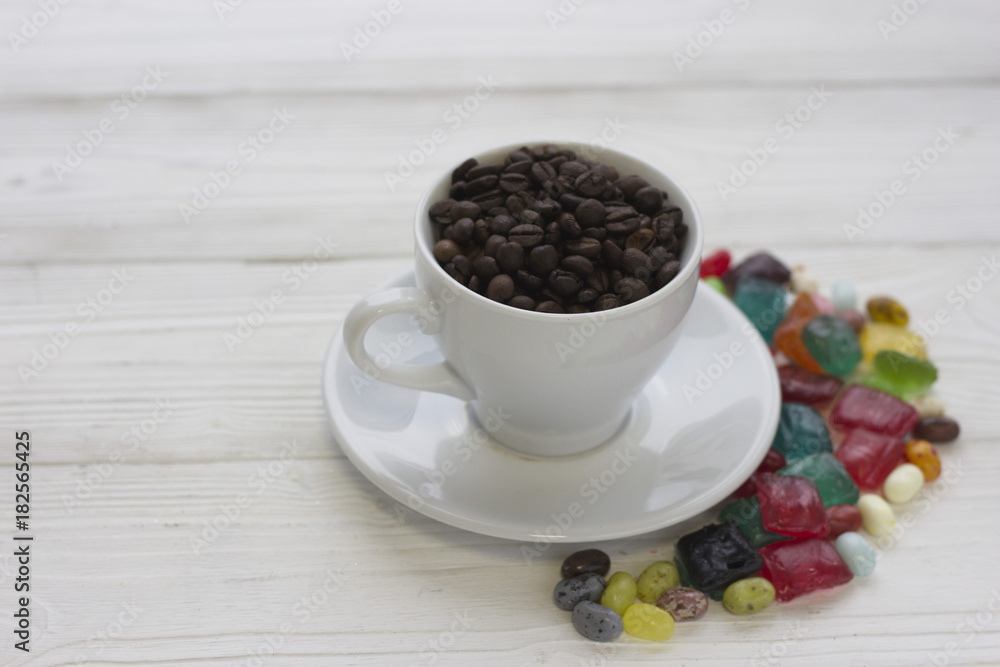 Festive still life with a cup of coffee with a saucer and spilled colored candies. The cup is filled with coffee beans. Close up. Light wooden background. Copy space Festive still life with a cup of coffee with a saucer and spilled colored candies. The cup is filled with coffee beans. Close up. Light wooden background. Copy space