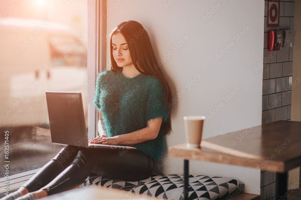 Fototapeta premium Beautiful brunette using laptop in cafe. Young attractive woman is making plans for the future, sitting in front of an open laptop computer in a cozy coffee shop.