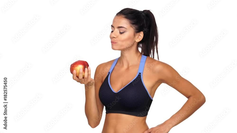 Pleased brunette sports woman eating apple and looking at the camera while holding arm on hip over white background Pleased brunette sports woman eating apple and looking at the camera while holding arm on hip over white background