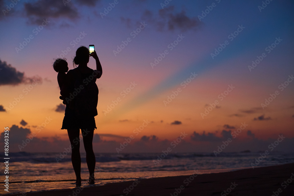 silhouette of young mother carrying child making photo of sunset by smartphone