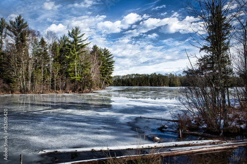 Tranquil winter lake and forest with reflection of trees and clouds on icy water