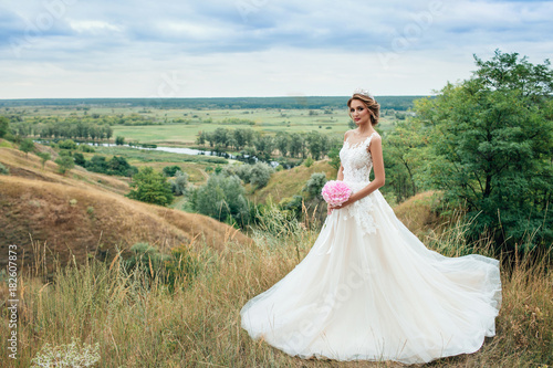 Beautiful young bride girl smiling, standing in wedding dress. Overlooking the beautiful green valley.