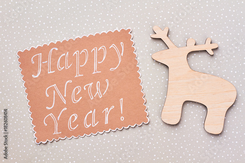 Happy New Year wooden background. Carved wooden deer and paper note with text Happy New Year on grey dotted background. New Year handmade craft.
