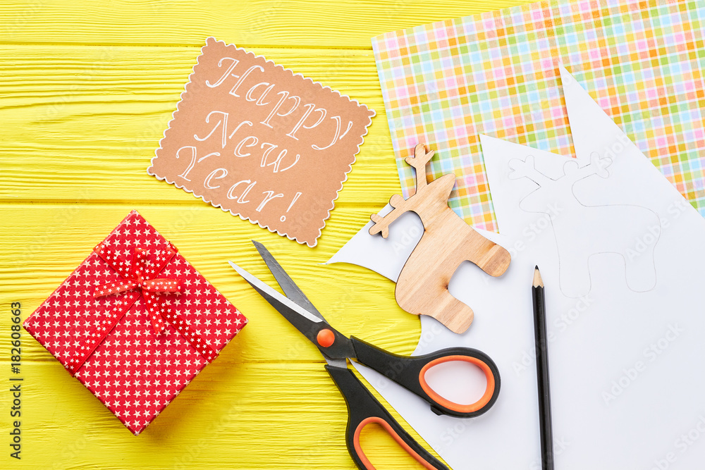 Fototapeta premium New Year handmade background, top view. Cut out wooden decoration deer, scissors, pencil, gift box, message on yellow wooden background. Happy New Year.