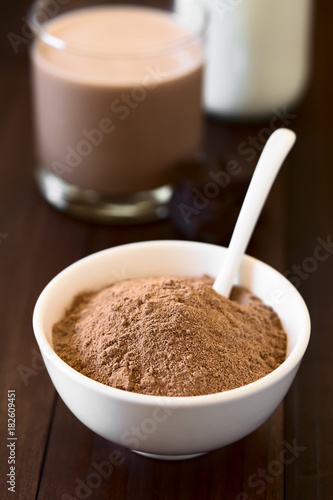 Cocoa or chocolate drink powder in bowl with spoon, chocolate drink and milk in the back, photographed with natural light (Selective Focus, Focus one third into the image)