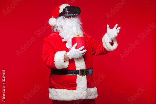 Santa Claus wearing virtual reality goggles and a red bucket with popcorn, on a red background. Christmas