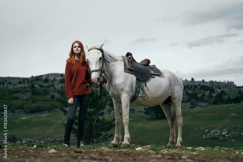 young red-haired girl is photographed with a white horse against the background of the countryside