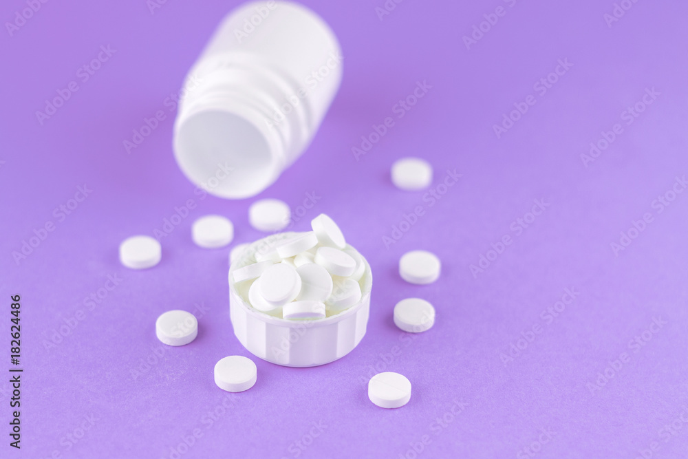 Close up white pill bottle with spilled out pills and capsules in cap on purple background with copy space. Focus on foreground, soft bokeh. Pharmacy drugstore concept Close up white pill bottle with spilled out pills and capsules in cap on purple background with copy space. Focus on foreground, soft bokeh. Pharmacy drugstore concept