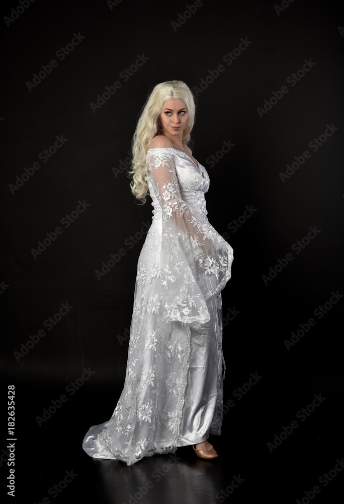 full length portrait of a blonde lady wearing beautiful lace gown, standing pose on black background.