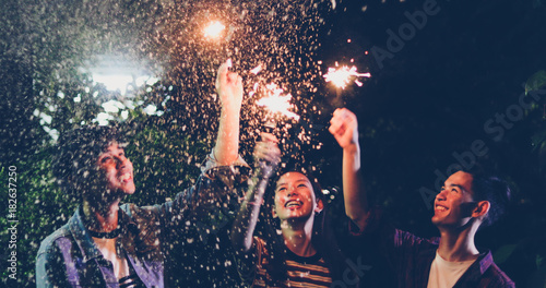 Asian group of friends having outdoor garden barbecue laughing with alcoholic beer drinks and showing group of friends having fun with sparklers on night ,soft focus
