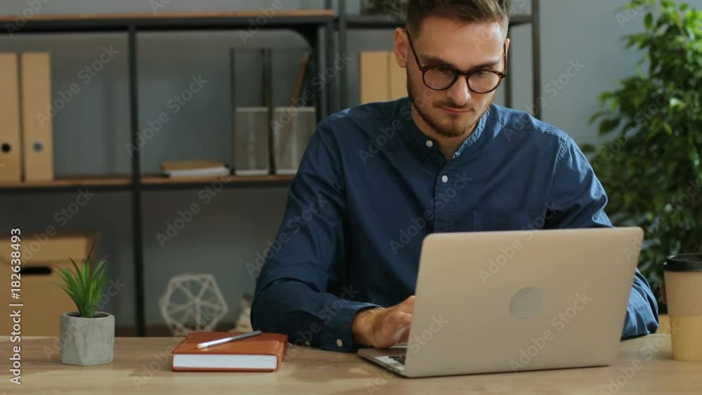 Portrait of serious young businessman in the glasses working on laptop computer in the modern office. Portrait of serious young businessman in the glasses working on laptop computer in the modern office.