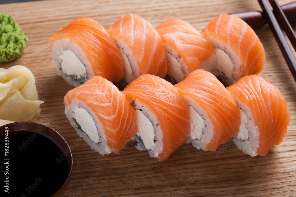 A classic Philadelphia roll with wasabi, ginger and soy sauce on a wooden Board. Salmon, Philadelphia cheese, cucumber, avocado. Japanese sushi on a dark stone background. Top view A classic Philadelphia roll with wasabi, ginger and soy sauce on a wooden Board. Salmon, Philadelphia cheese, cucumber, avocado. Japanese sushi on a dark stone background. Top view