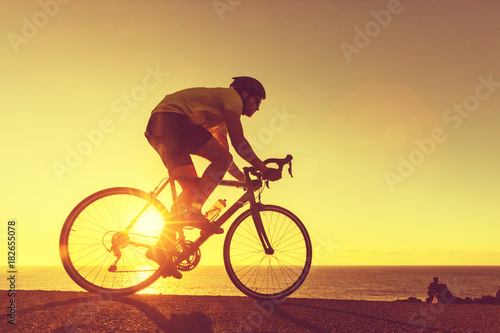 Fototapeta Naklejka Na Ścianę i Meble -  Road bike cyclist man biking on professional racing bike. Sports fitness triathlon athlete riding bike on road sunset with sun flare. Active healthy sports lifestyle athlete cycling.