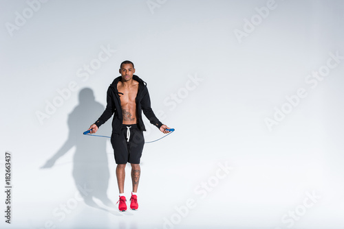young african american sportsman exercising with skipping rope and looking at camera on grey
