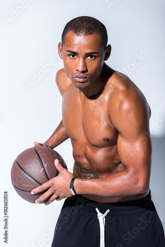 handsome african american man holding basketball ball and looking at camera on grey