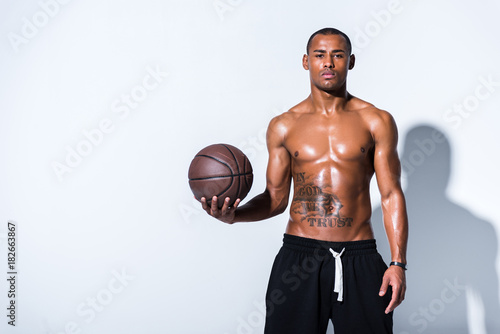 athletic shirtless african american man with basketball ball looking at camera on grey