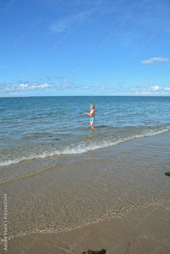 Kleiner blonder Junge mit blauer Badehose steht mit einem Kescher am Sandstrand der Nordsee, mit viel Strand und blauem Himmel