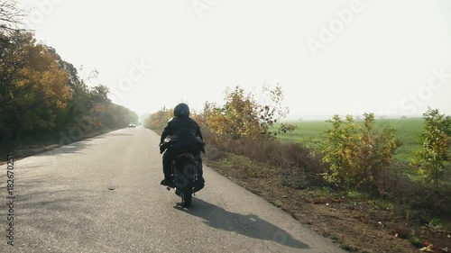 Back view of a man in black helmet and leather jacket riding motorcycle on a asphalt road on sunny day in autumn. Trees with brown and yellow leaves around the road photo