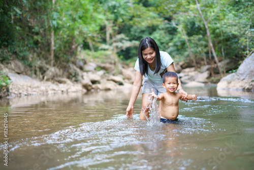 Fototapeta Naklejka Na Ścianę i Meble -  Asian mother in blue shirt with DIY chest pocket playing water with her son about 1 year and 9 months at  waterfall