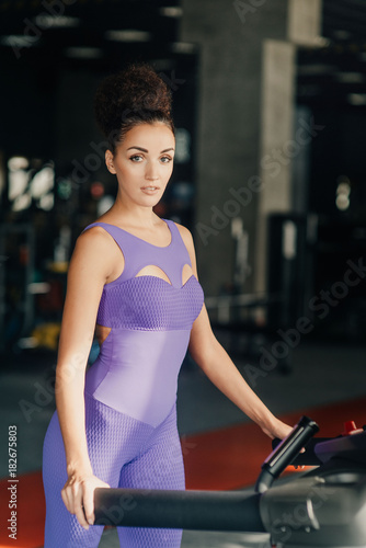 Portrait of alone young adult happy woman with long flying hair running on treadmill in gym wearing in sexy violet suit. Slim sport body.