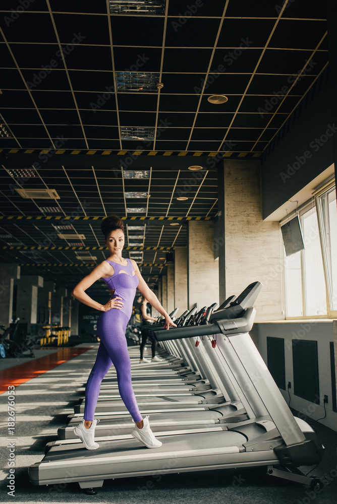 Running together. Side view of young beautiful woman looking in camera and posing before running on treadmill at gym Running together. Side view of young beautiful woman looking in camera and posing before running on treadmill at gym