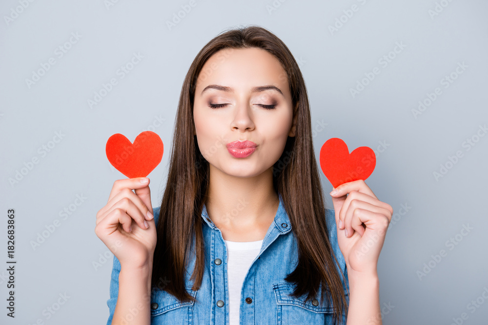 Close up portrait of brunette girl, holding two small red hearts in her raised hands, blowing kiss to the camera with closed eyes, dreaming about her lover while standing over grey background Close up portrait of brunette girl, holding two small red hearts in her raised hands, blowing kiss to the camera with closed eyes, dreaming about her lover while standing over grey background