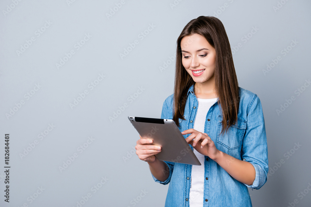 Close up portrait of smiling successful woman in casual outfit looking at her tablet and typing sms through 3g internet while standing over grey background Close up portrait of smiling successful woman in casual outfit looking at her tablet and typing sms through 3g internet while standing over grey background