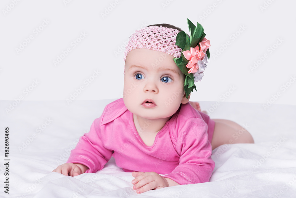 cute little baby girl with bow flower on her head. child with big blue eyes lies on his belly on soft blanket, isolated over gray background cute little baby girl with bow flower on her head. child with big blue eyes lies on his belly on soft blanket, isolated over gray background