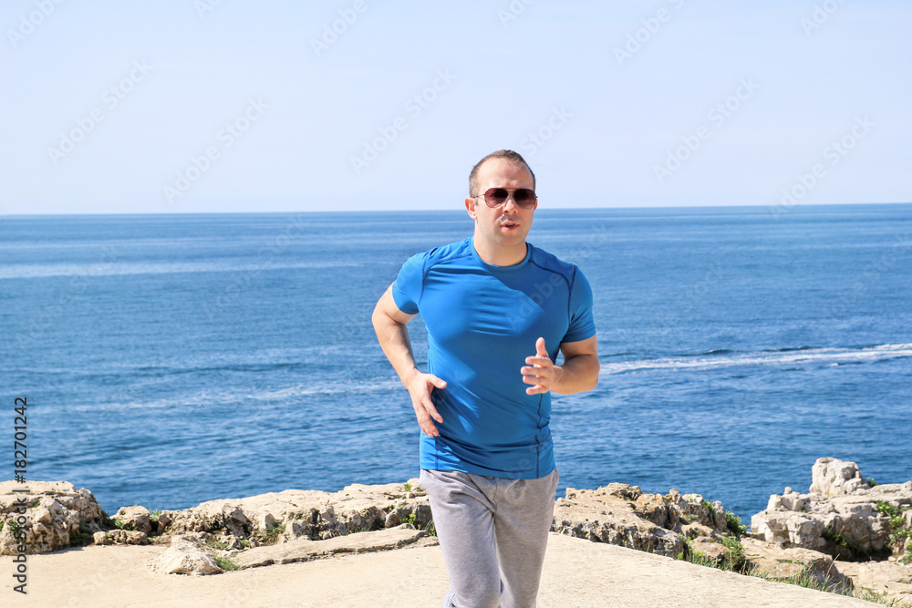 Fit young man jogging on a running trail along seashore. Recreational fitness athlete in sportswear enjoys physical activities on a summer day in a beautiful natural environment. Healthy lifestyle.