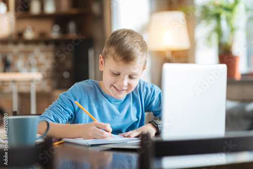 School assignment. Positive nice smart boy sitting at the table and doing his home task while preparing for school