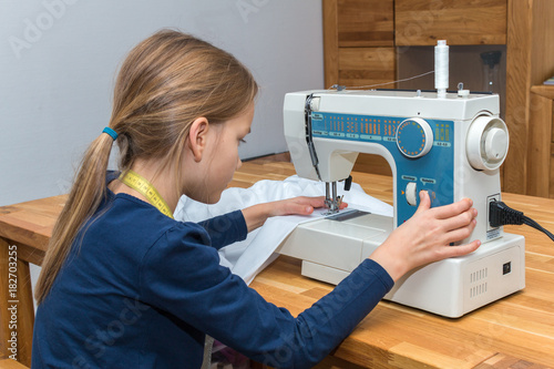 Ein Mädchen sitz am Tisch und näht mit der Nähmaschine einen weißen Stoff um