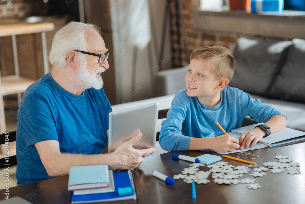 School lessons. Cheerful positive smart man holding a tablet and looking at his grandson while helping him with his school lessons School lessons. Cheerful positive smart man holding a tablet and looking at his grandson while helping him with his school lessons