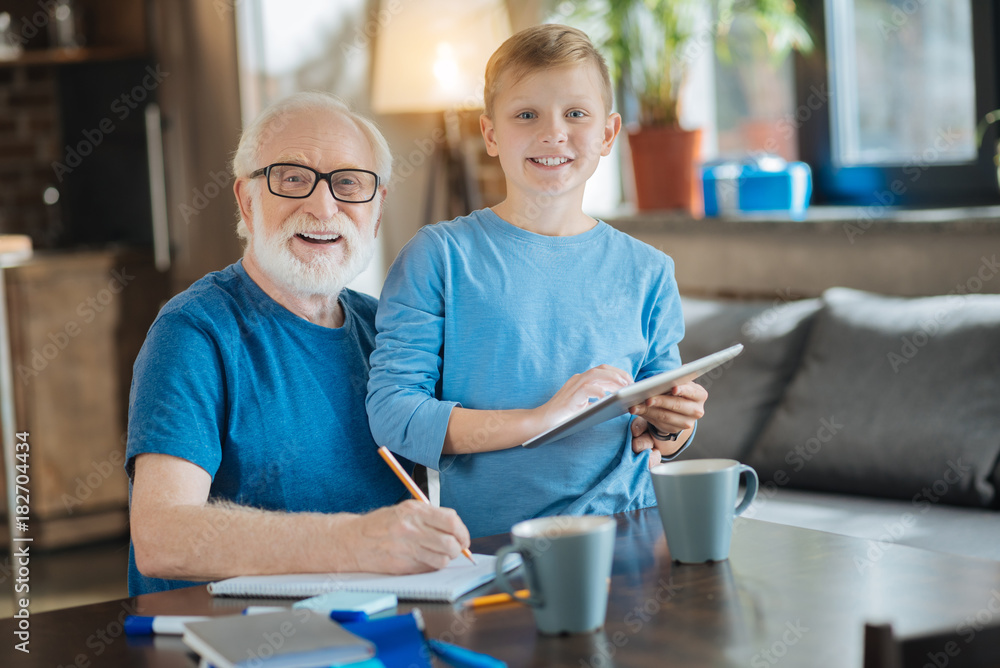 Fototapeta premium Modern igen. Nice cheerful delighted boy having a tablet in his hands and smiling while standing together with grandfather