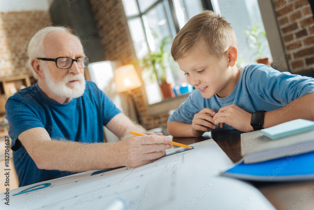 Future engineer. Nice happy curious boy sitting at the table and smiling while looking at his grandfathers drawing Future engineer. Nice happy curious boy sitting at the table and smiling while looking at his grandfathers drawing