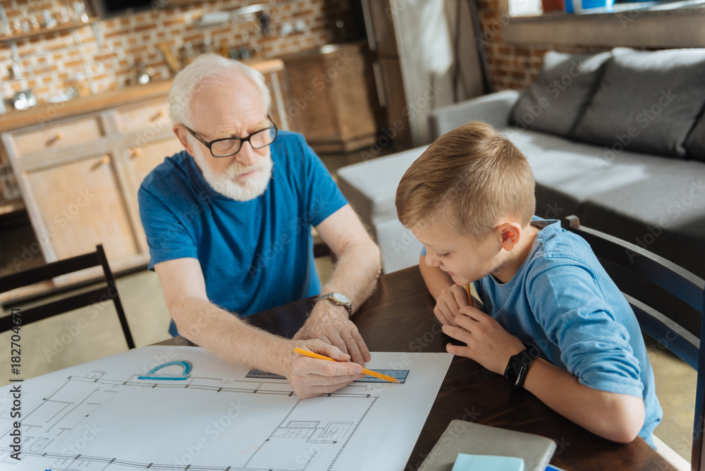 Professional scheme. Positive nice smart boy looking at the drawing and being interested while watching his grandfather working Professional scheme. Positive nice smart boy looking at the drawing and being interested while watching his grandfather working