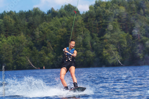 Young pretty slim brunette woman riding wakeboard on wave of motorboat in summer lake