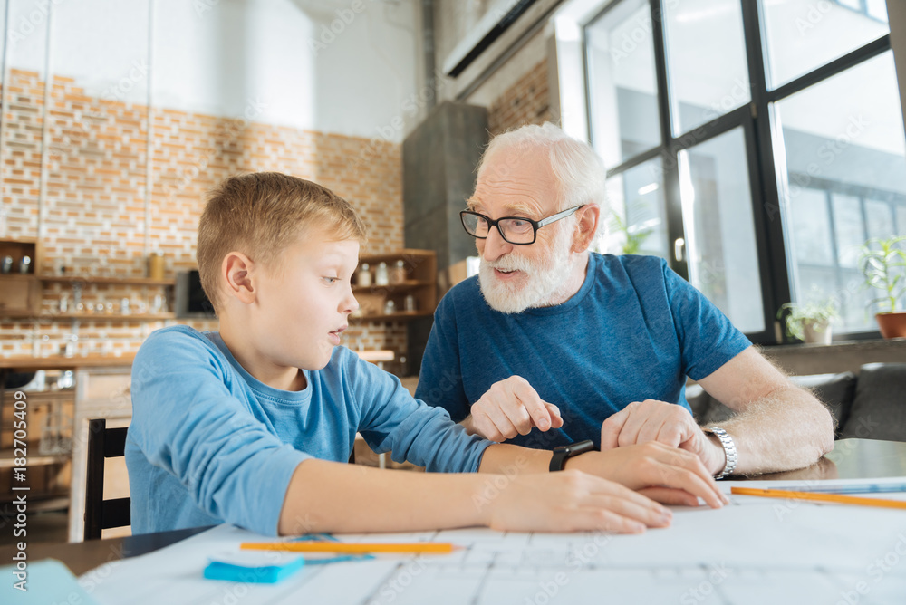 What is this. Joyful curious aged man looking at his grandson and pointing at the smartwatch while wondering what it is What is this. Joyful curious aged man looking at his grandson and pointing at the smartwatch while wondering what it is