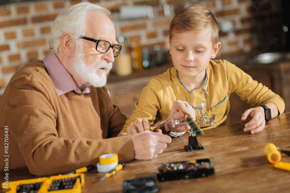 Important skills. Nice pleasant smart grandfather and grandson sitting together and the table and fixing objects while enjoying this activity Important skills. Nice pleasant smart grandfather and grandson sitting together and the table and fixing objects while enjoying this activity