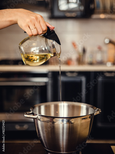 Female hand pouring olive oil in glass jag into a big metal bowl for making a dough. Home cooking on kitchen.