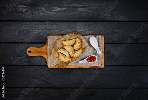 Hot fried chebureks with sour cream and sauce on a wooden board. Top view. Black wooden background