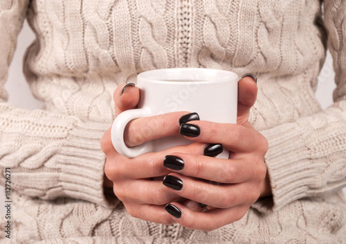 Girl in sweater holding cup of tea in her hands with black nails, close up