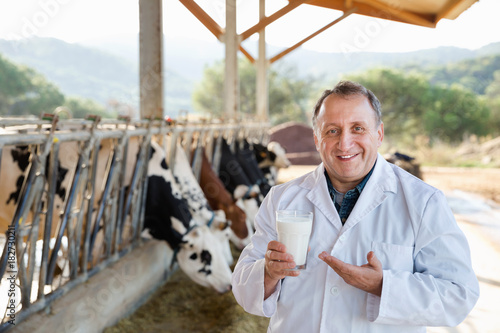 Male farmer is holding glass of cow milk at the cow farm.