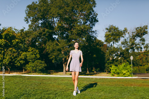 Young pretty girl with long brown hair dressed in light clothes staying on green lawn grass in the park on trees background. Summer sunny time.