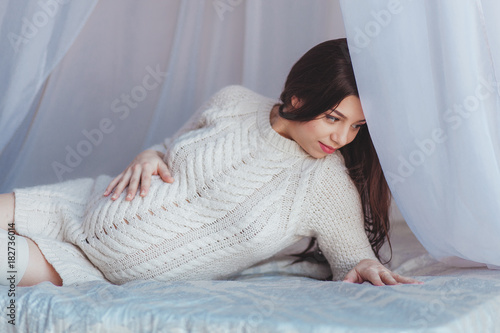 Beautiful Pregnant Woman lying in bed with canopy. Wears white knitted pullover. Maternity concept photo