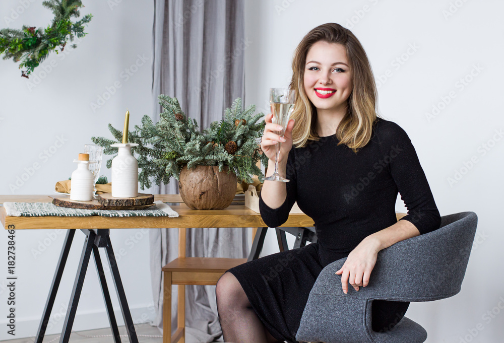Young happy beautiful woman is sitting with glass of champagne  near the table and celebrating Christmas and New Year.