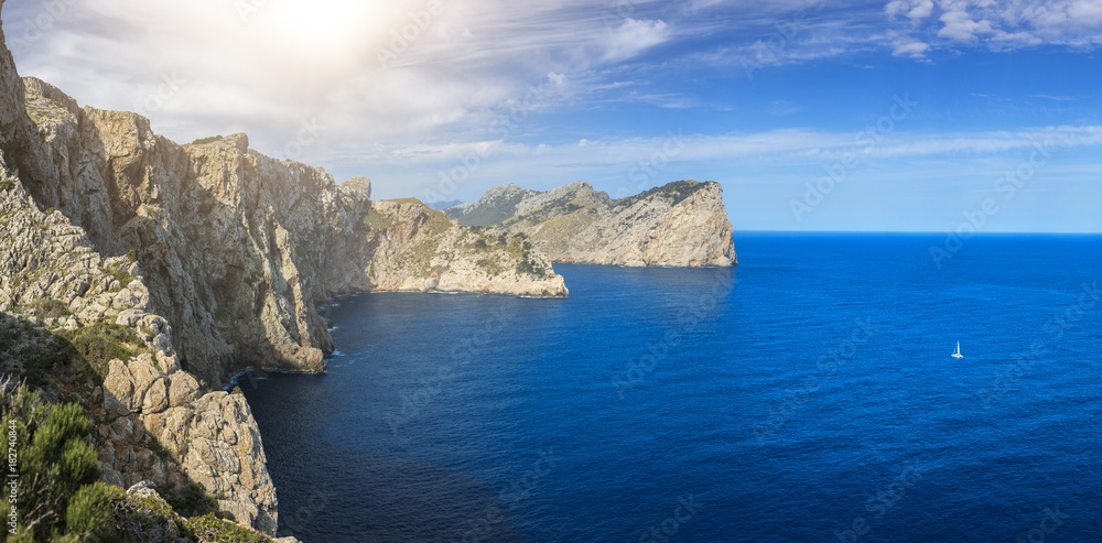 A panoramic picture of the cliffs on the Pollença coastline and a single white sailboat sailing on the calm sea on a nice sunny day in Mallorca