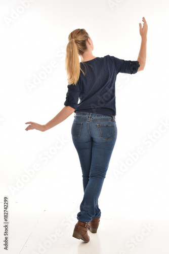 Full length portrait of a girl wearing simple blue shirt and jeans, standing pose facing away on a white background.