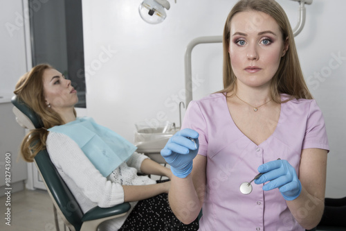 Young dental doctor girl holding a medical instrument against the background of a dental office. In the chair sits a patient