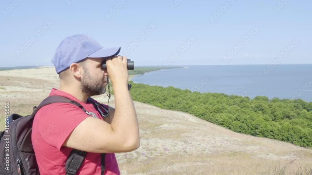Travel man looking at wildlife with binoculars. Male on vacation looking at nature landscape. Hiker with a backpack is looking through binoculars at sea, at beautiful landscape. Mountain. Beach. Travel man looking at wildlife with binoculars. Male on vacation looking at nature landscape. Hiker with a backpack is looking through binoculars at sea, at beautiful landscape. Mountain. Beach.