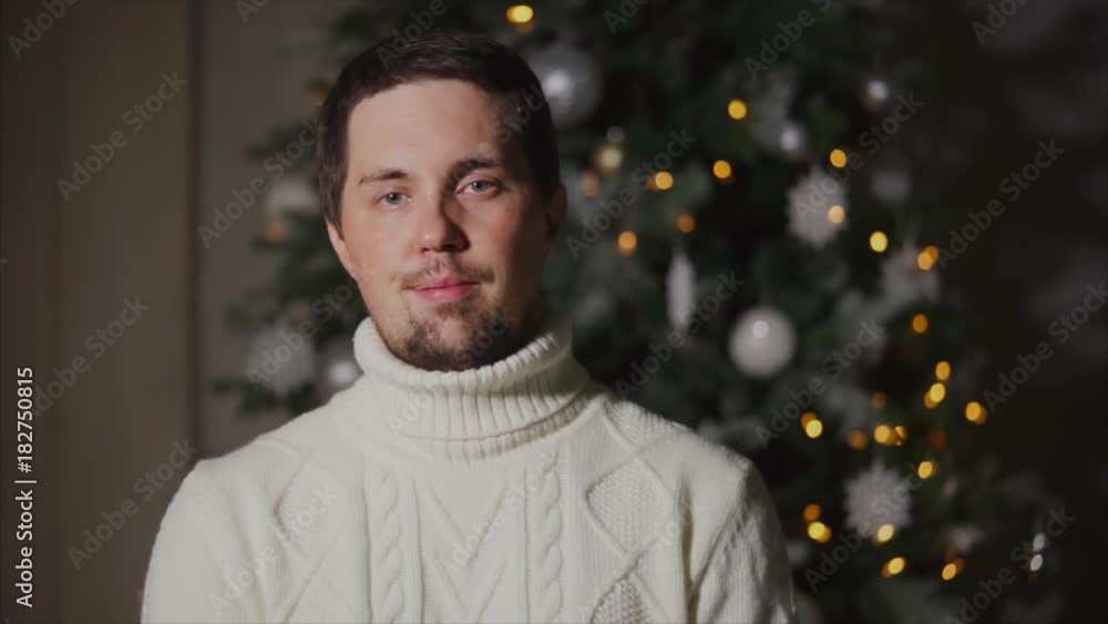 portrait of a young man in a winter sweater who stands near the Christmas tree, a Christmas tree decorated with toys and lights, calm people ready to celebrate with friends portrait of a young man in a winter sweater who stands near the Christmas tree, a Christmas tree decorated with toys and lights, calm people ready to celebrate with friends